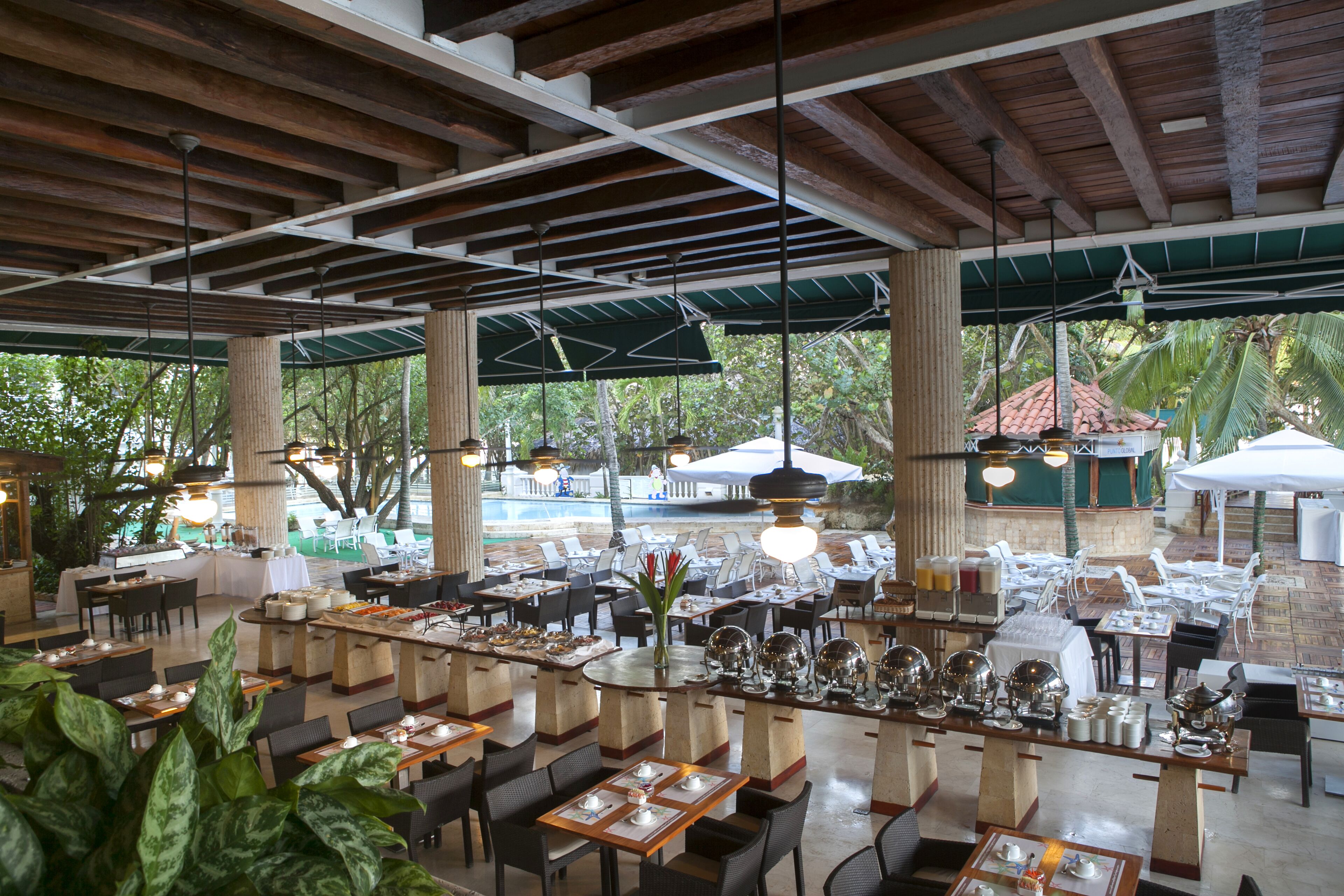 Un vaste espace extérieur aménagé pour les repas, avec de nombreuses tables et chaises, de grands parasols et une vue sur une piscine et des palmiers.