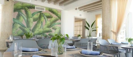 A dining area with tables set for service, a large tropical leaf mural on the wall, and potted plants.