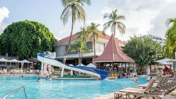 Una piscina con sillones de descanso, un tobogán de agua, palmeras y un edificio con techo rojo.
