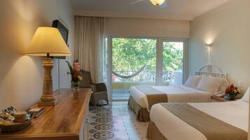 Quarto de hotel com duas camas, uma escrivaninha de madeira com luminária, uma cadeira, varanda com rede e vista para o verde.