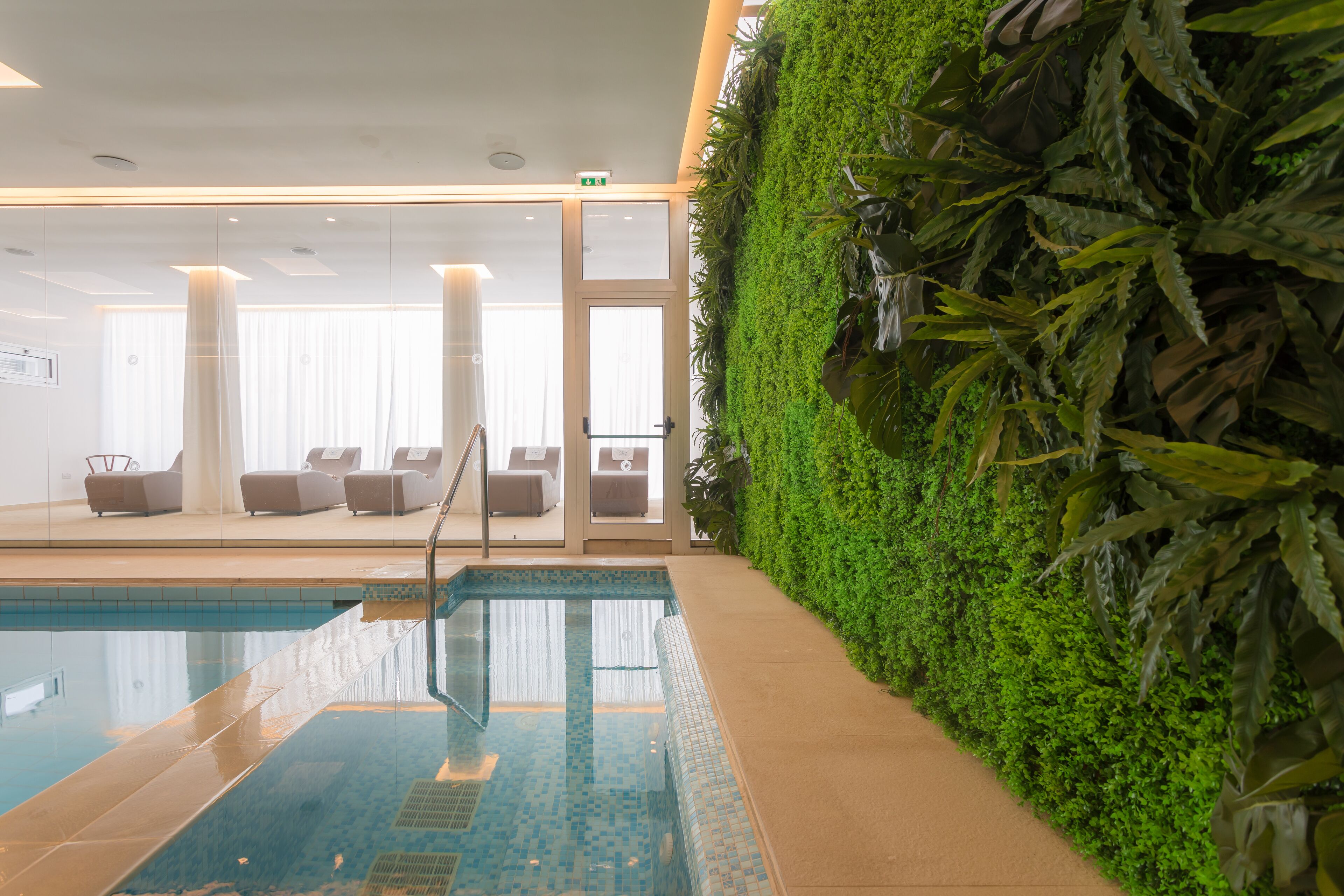 A swimming pool with a glass wall, a seating area with chairs, and a green wall with plants.