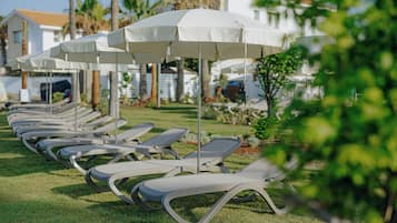 A row of white sun loungers with matching umbrellas, arranged on a grassy lawn with a building in the background.