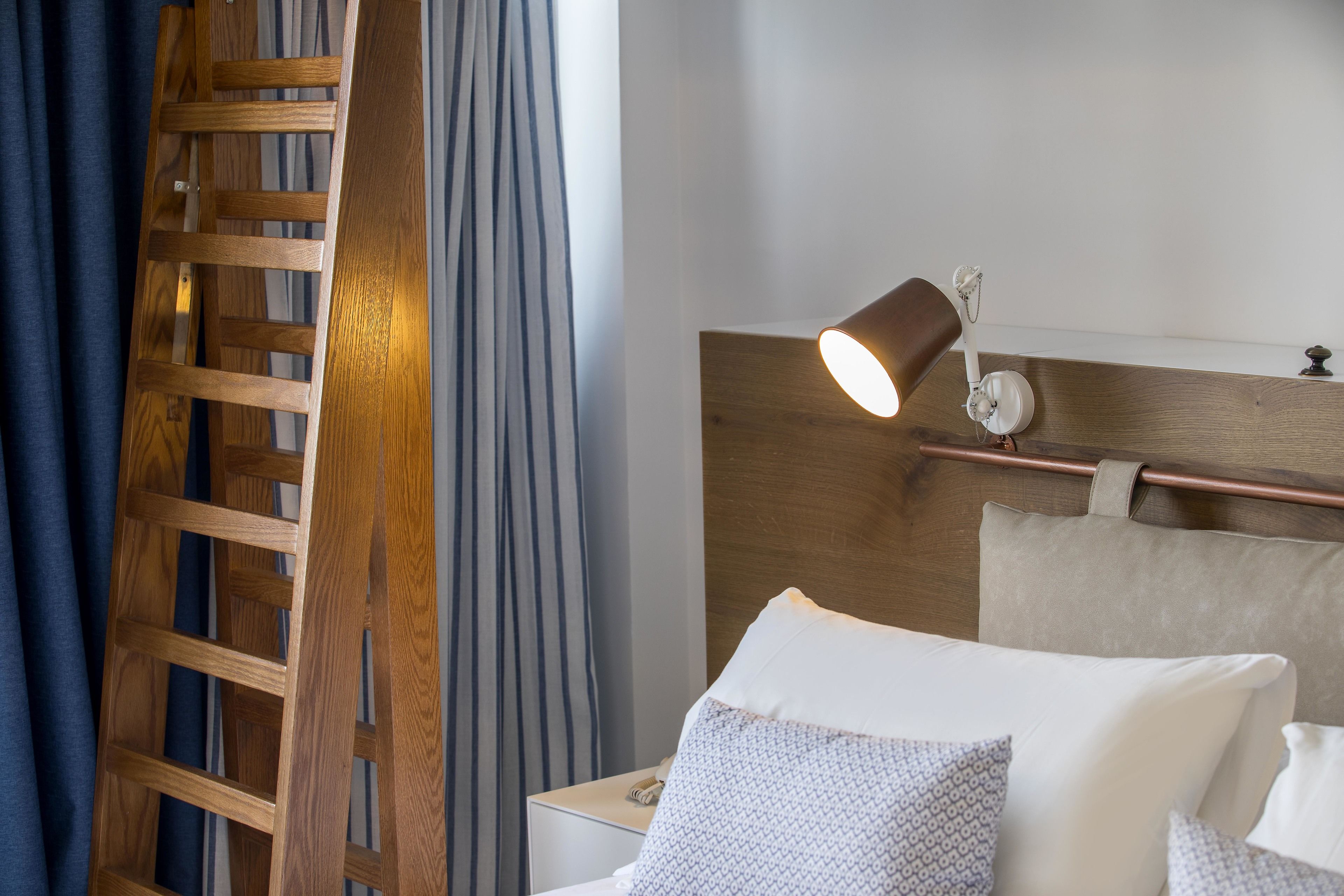 A hotel room with a wooden ladder, a bedside lamp, a bed with pillows, and a nightstand.