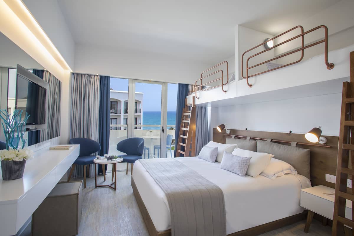 A hotel room with a bed, a desk, a chair, a small table, a mirror, and a view of the sea.