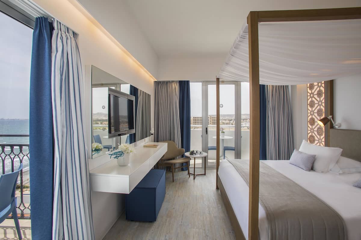 A hotel room with a bed, a desk, a chair, a small table, and a balcony with curtains.