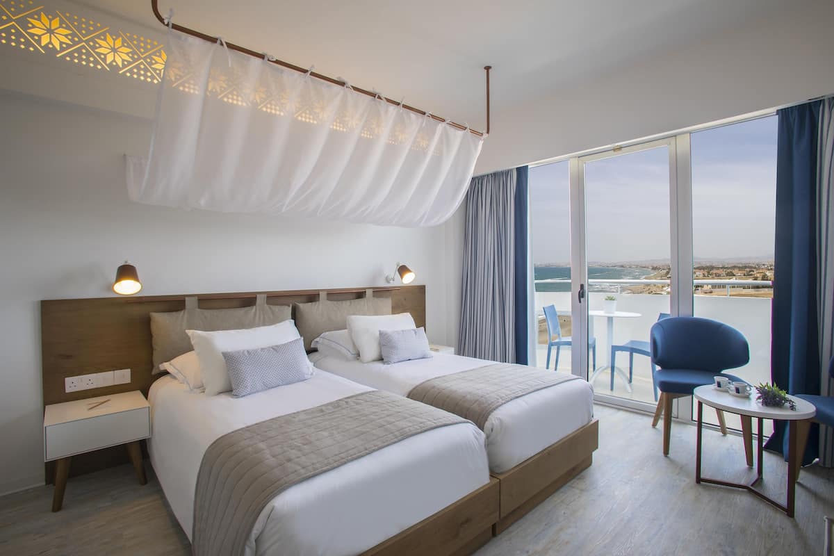 A hotel room with two beds, a small table, a chair, and a balcony with a view.