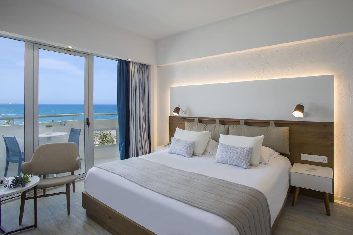 A hotel room with a large bed, a bedside table, a chair, and a view of the ocean.
