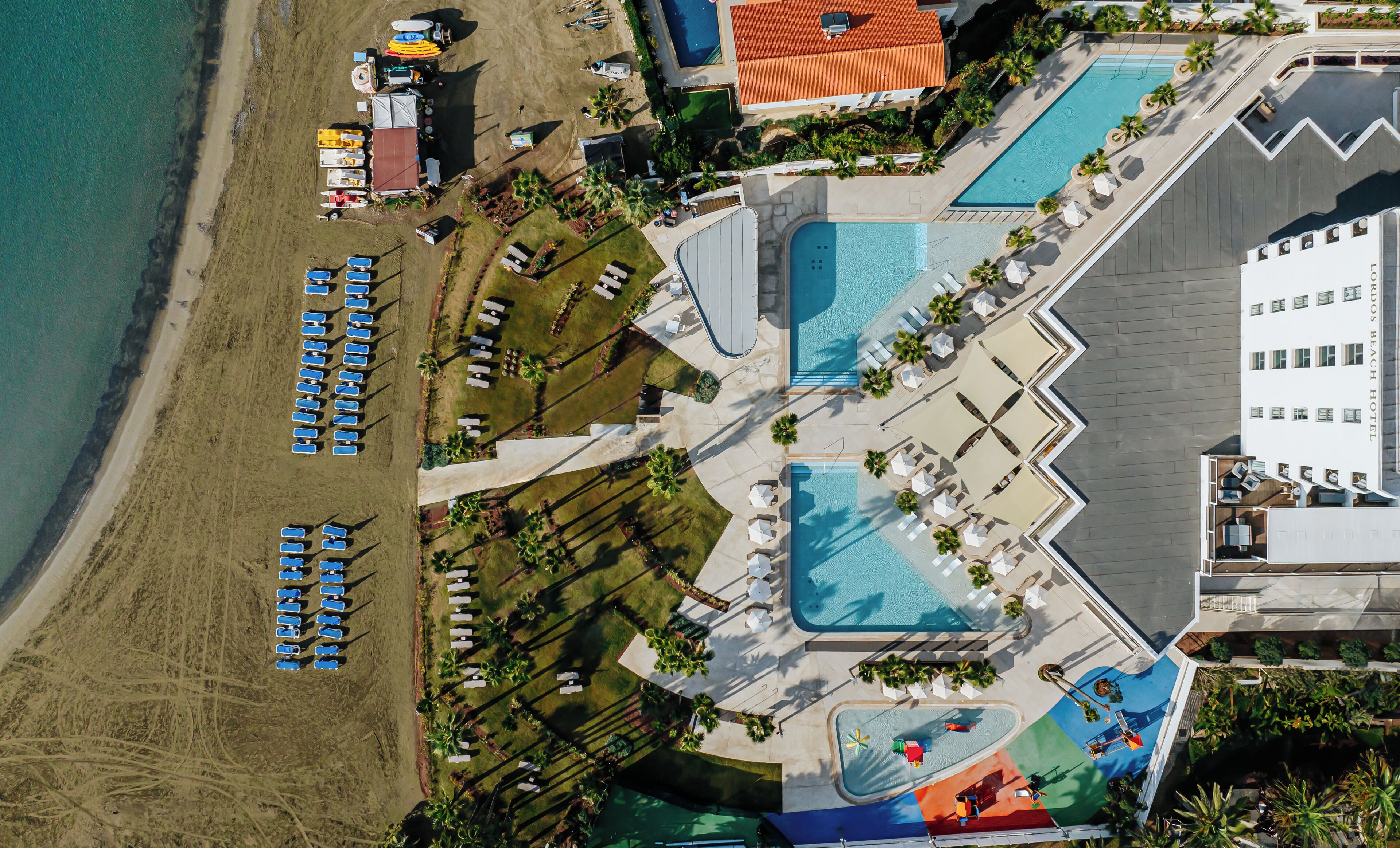 Ett flygbildsfoto av ett semesterområde med flera pooler, strandhytter och en stor byggnad med ett rött tak.