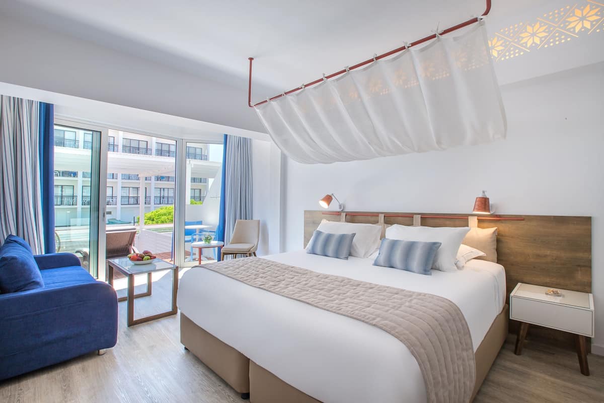 A modern hotel room with a large bed, a blue sofa, a small table with fruit, and a view of the city through large windows.