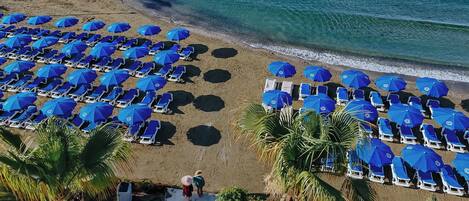 An aerial view of a beach with rows of blue umbrellas and white lounge chairs, palm trees, and a clear view of the ocean.