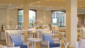 A spacious, well-lit lounge area with white furniture, including sofas and chairs, arranged around tables.