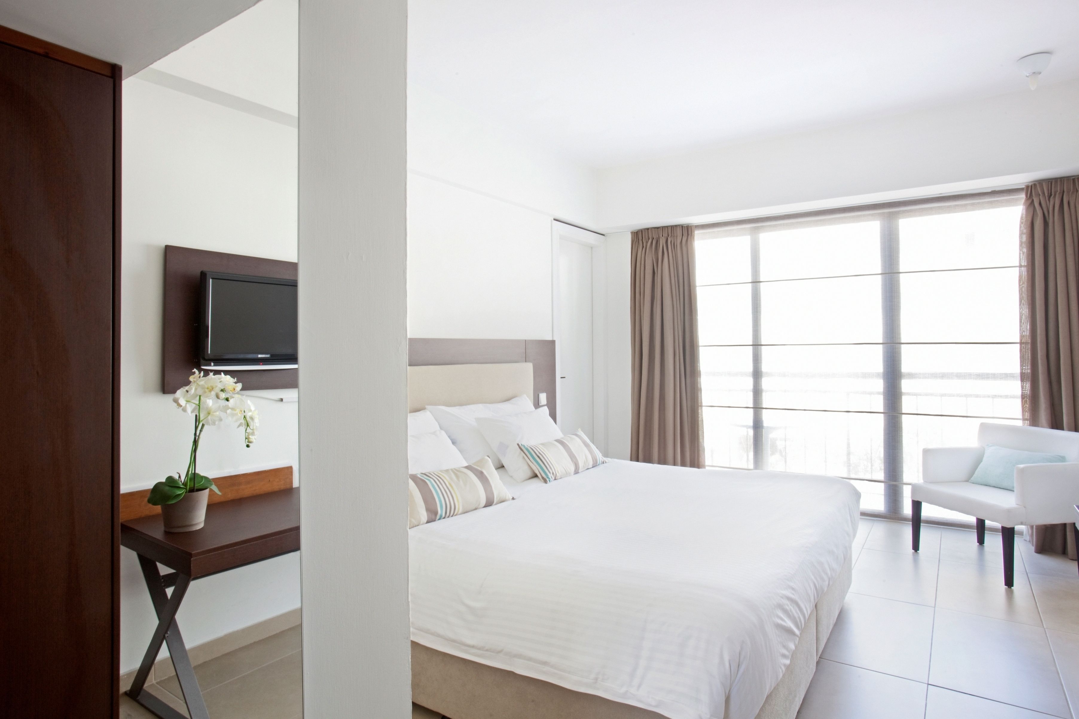 Quarto de hotel com uma cama grande, uma escrivaninha com planta em vaso, uma TV na parede e vista para o exterior através de amplas janelas.