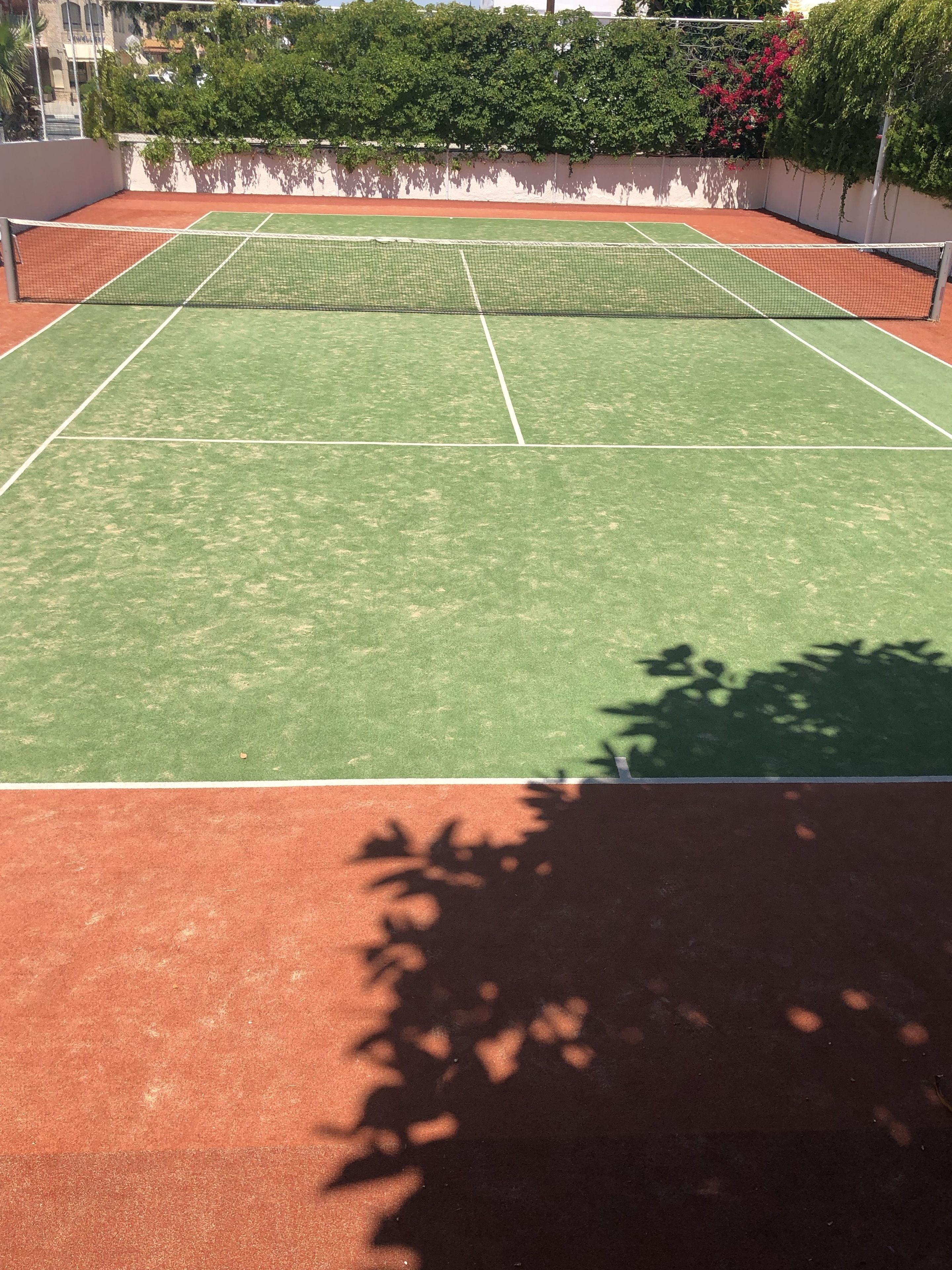 A tennis court with a net, surrounded by a red surface and greenery.