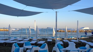 A beachside lounge area with white lounge chairs and blue cushions, umbrellas providing shade, and a view of the sea and rocky shoreline.