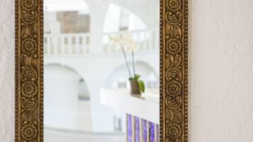 Un miroir doté d’un cadre doré orné, reflétant un intérieur moderne avec un mur blanc et une étagère décorative.