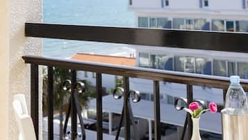 Un balcón con una mesa blanca, una silla de mimbre, un florero con flores y una botella. Se ve un edificio y el océano.