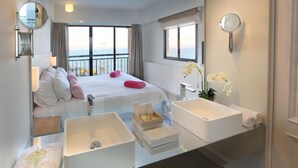 A hotel room with a large bed, two white sinks, a mirror, and a view of the sea.