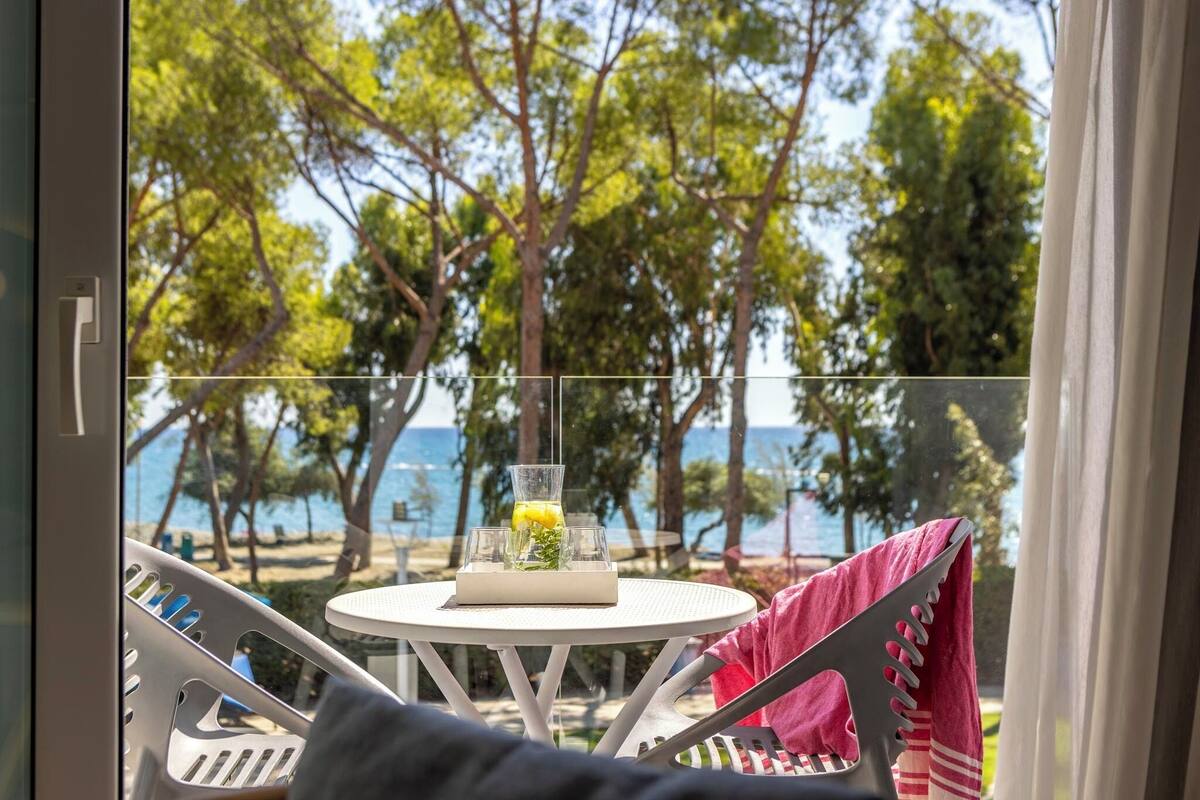 A balcony with a table set for two, overlooking a beach with trees and the sea.