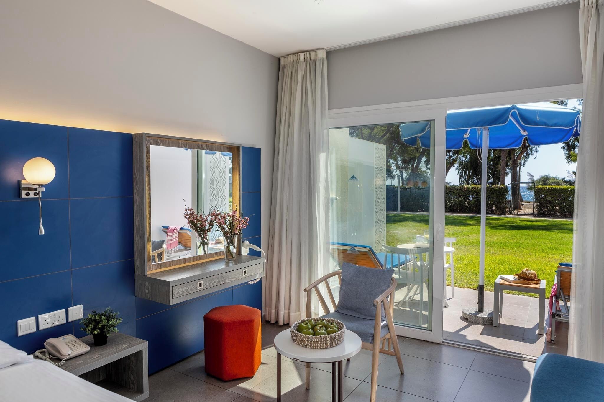 Habitación de hotel con un amplio ventanal corredera que da a un balcón, una cama, una mesita con una cesta, una silla y una mesita de noche con un teléfono.
