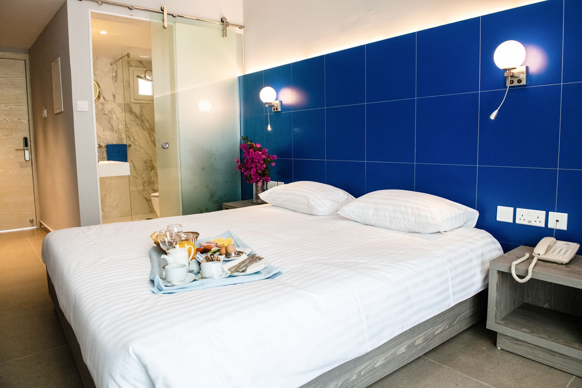 A hotel room with a bed, a bedside table with a phone, and a tray with breakfast items.
