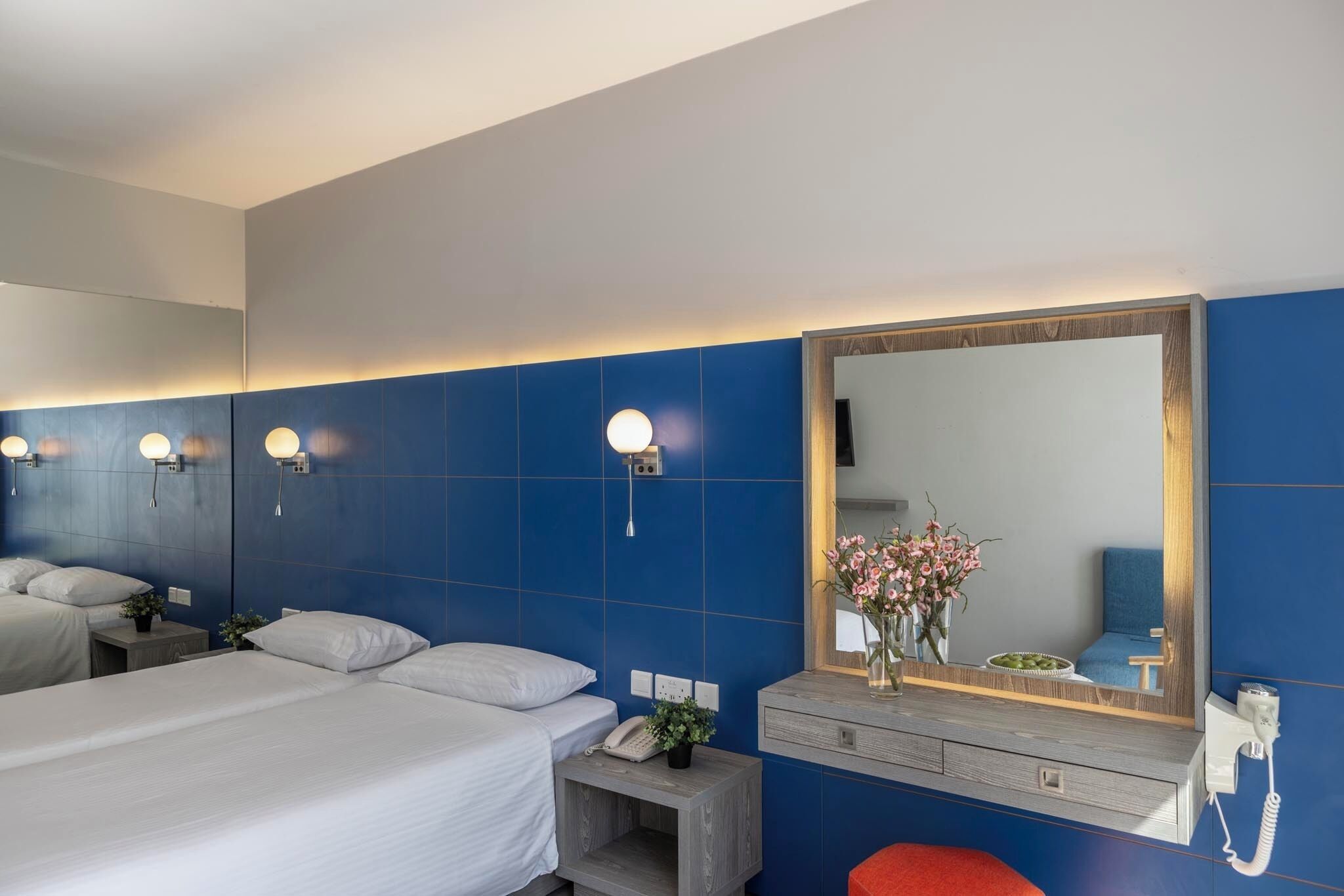 A hotel room with a large bed, a bedside table with a phone and a small plant, a mirror with a wooden frame, and a blue tiled wall.
