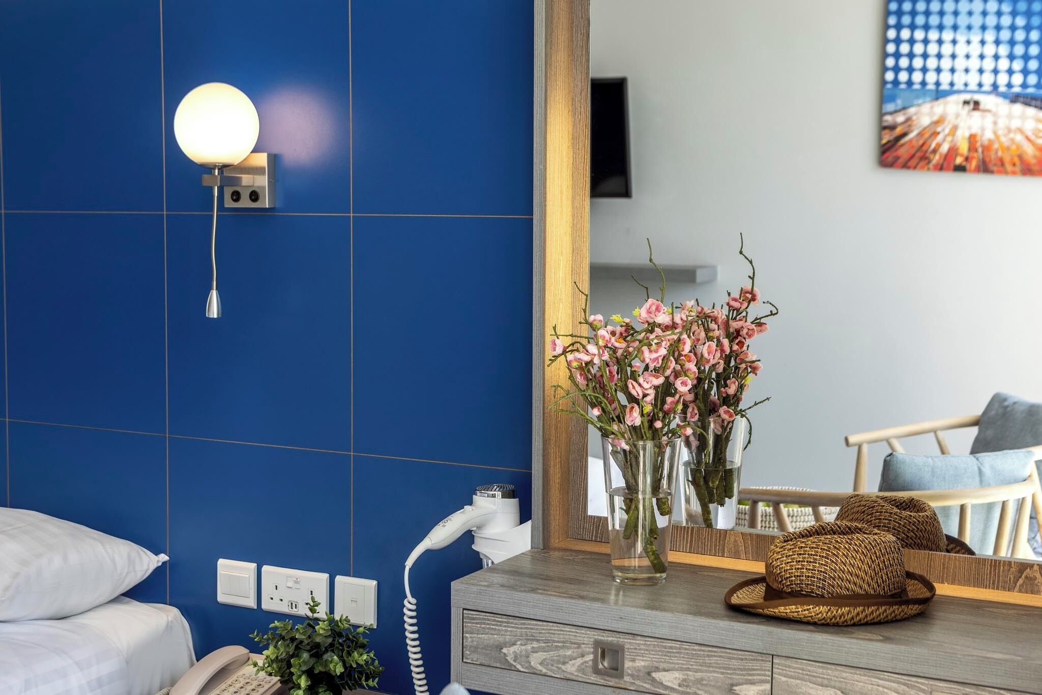 Ein Hotelzimmer mit blauer Fliesenwand, einem Nachttischtelefon, einem Spiegel, einer Vase mit rosa Blumen und einem Holznachtisch mit einem gewebten Korb.