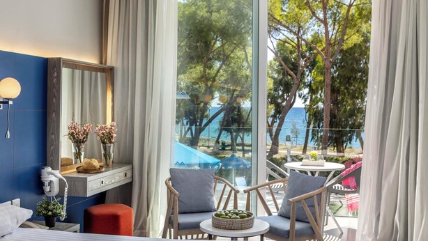 A hotel room with a large window, a desk with a mirror, two chairs, a small table with a bowl of fruit, and a bed. - Park Beach Hotel (Limassol)