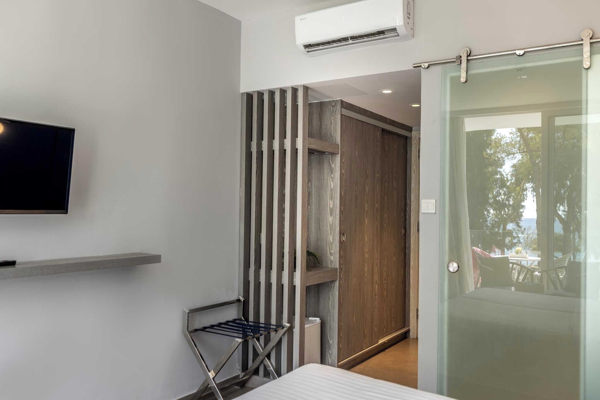 Una habitación de hotel moderna con televisión de pantalla plana, un armario de madera, una puerta de vidrio que da a un balcón y una pequeña zona de estar.