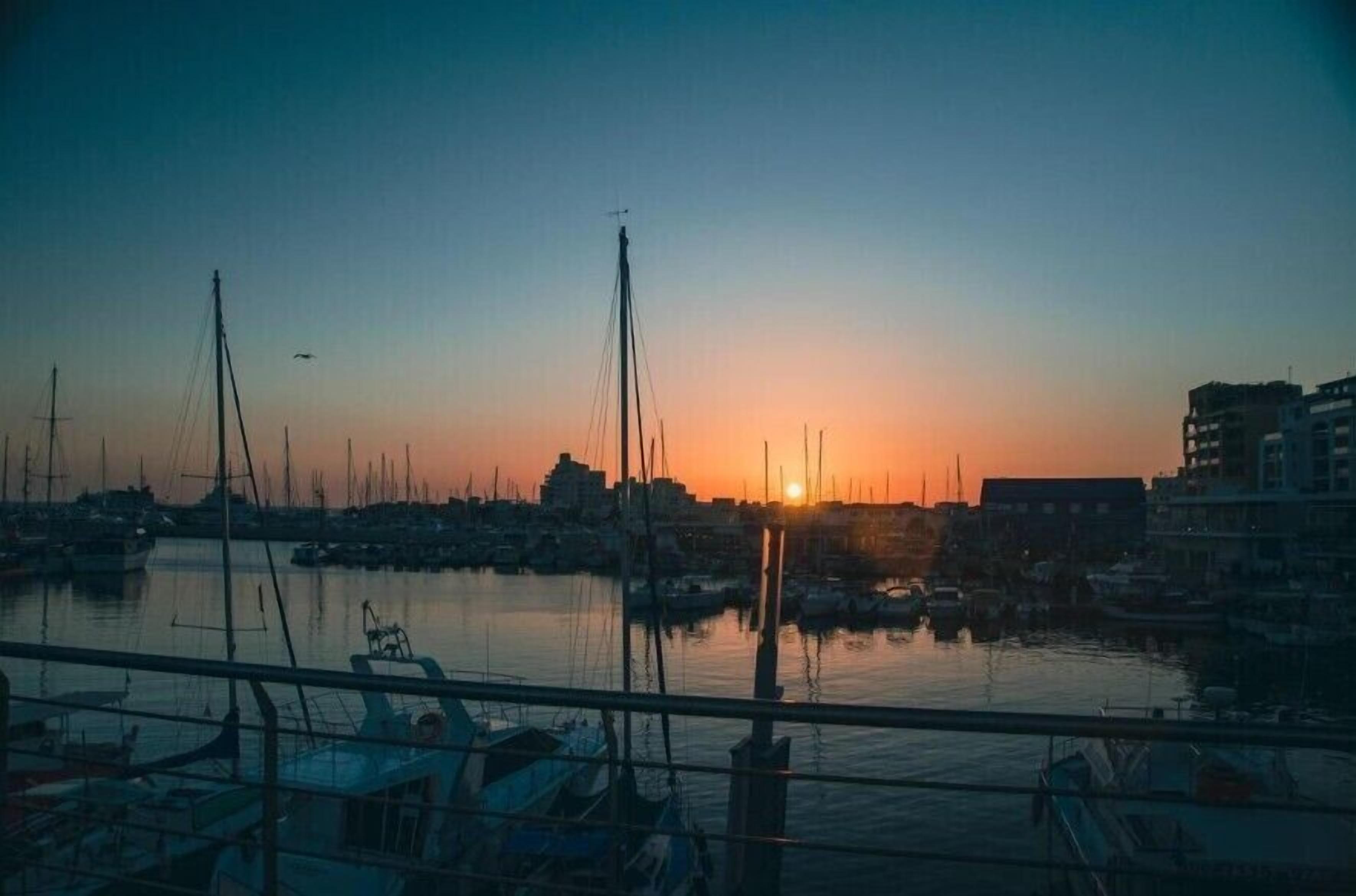 A marina at sunset with boats docked, a clear sky, and buildings in the background.