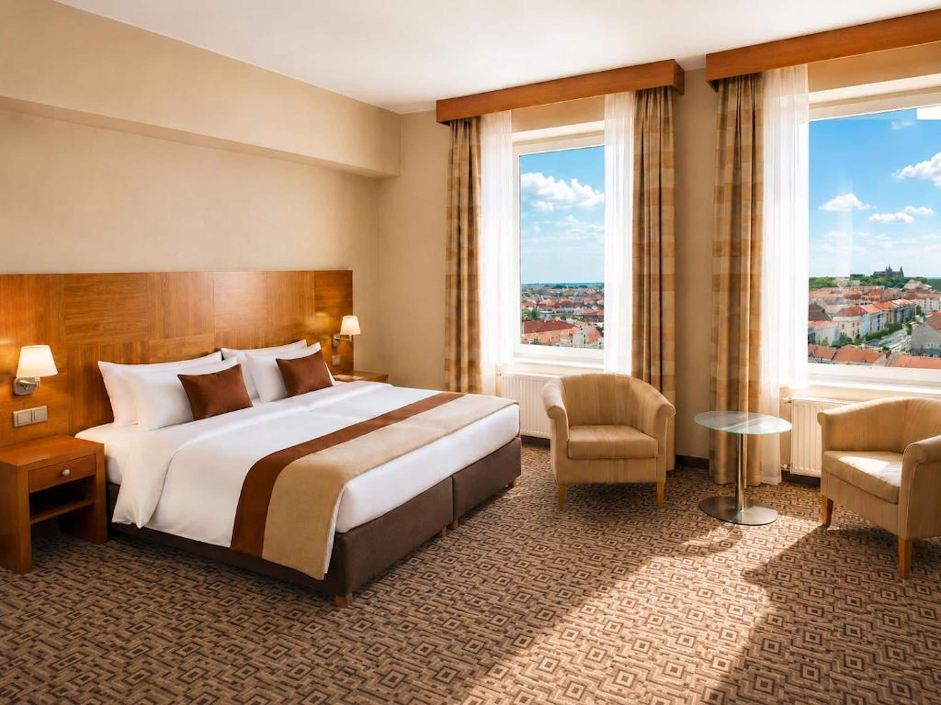 A hotel room with a large bed, two bedside tables, a chair, a small round table, and a view of a cityscape through large windows.
