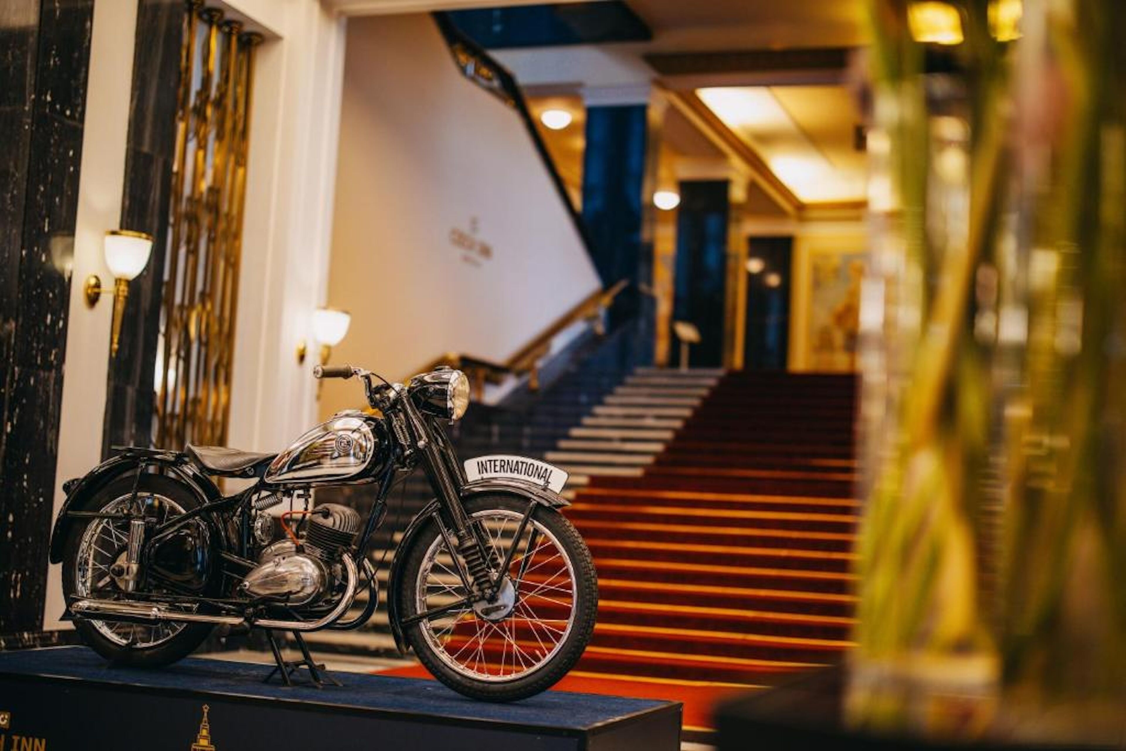 Una motocicleta antigua estacionada en una plataforma en el vestíbulo de un hotel, con una escalera al fondo.