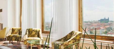 Una habitación amplia con un gran ventanal que ofrece vistas a la ciudad, dos sillones, una mesita y un ventanal de piso a techo con cortinas translúcidas.