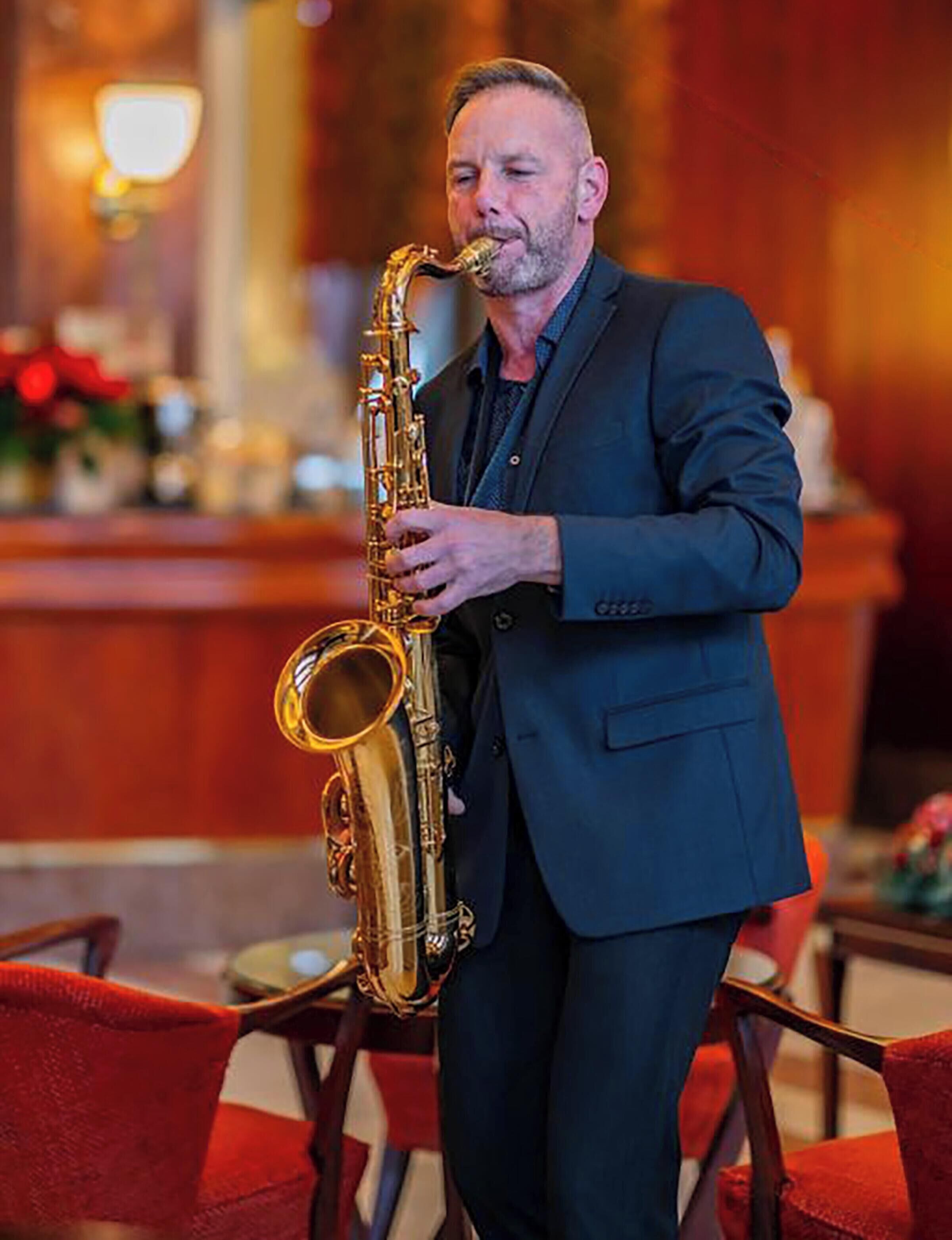 A man playing a saxophone in a warmly lit room with red chairs and a bar area in the background.