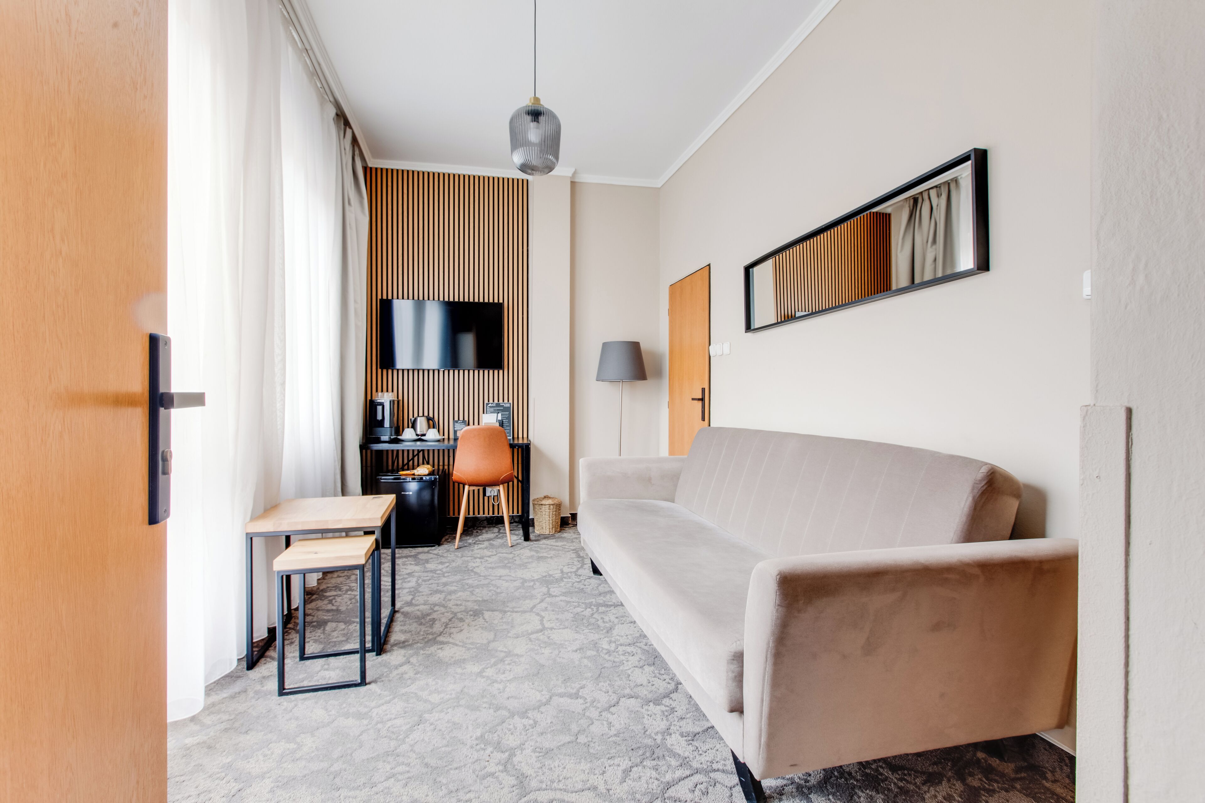 A modern hotel room with a sofa, a desk with a chair, a flat-screen TV, a lamp, and a framed picture on the wall.