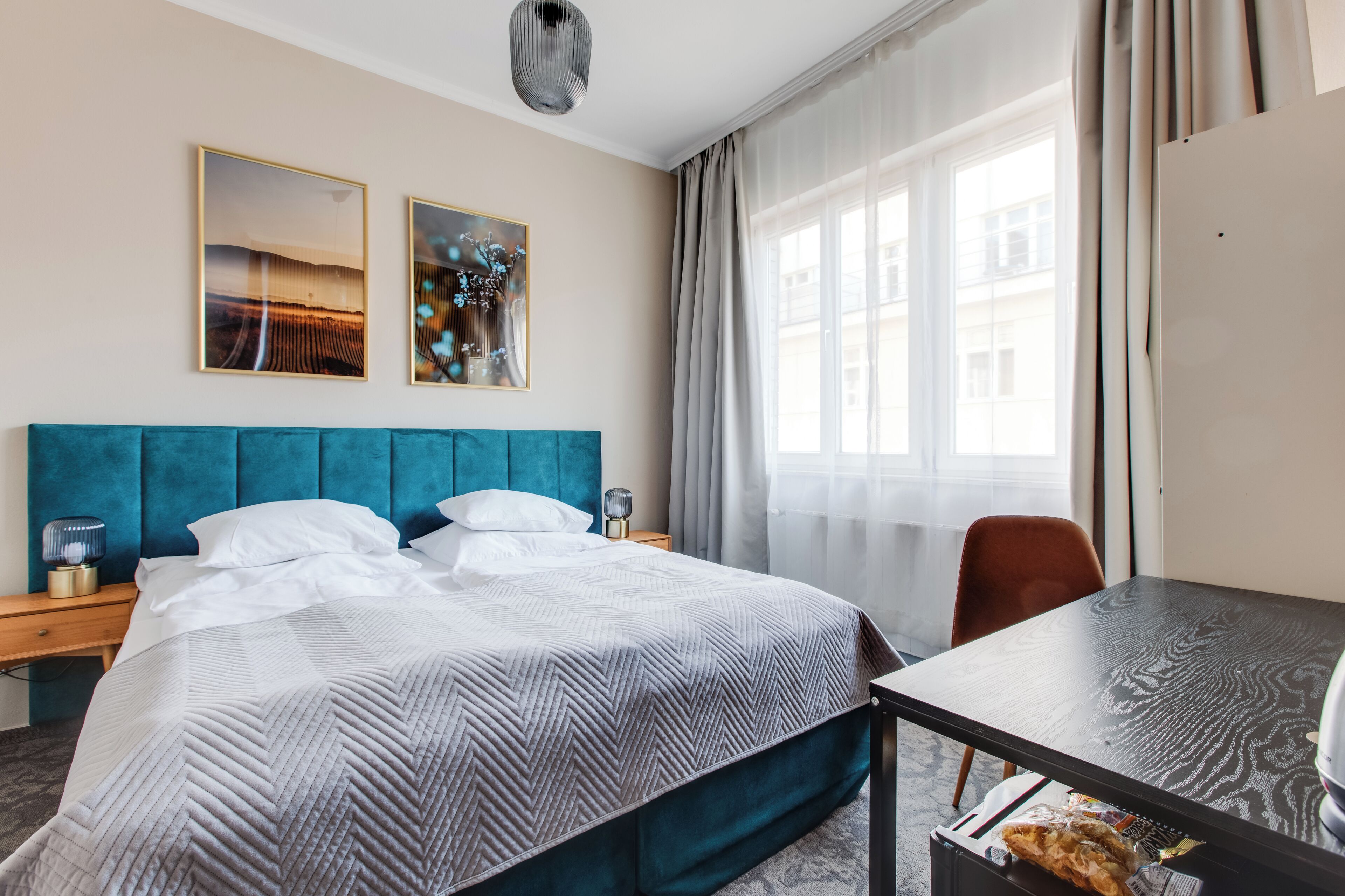 A hotel room with a large bed, a desk, and a chair. There are two paintings on the wall and a window with curtains.