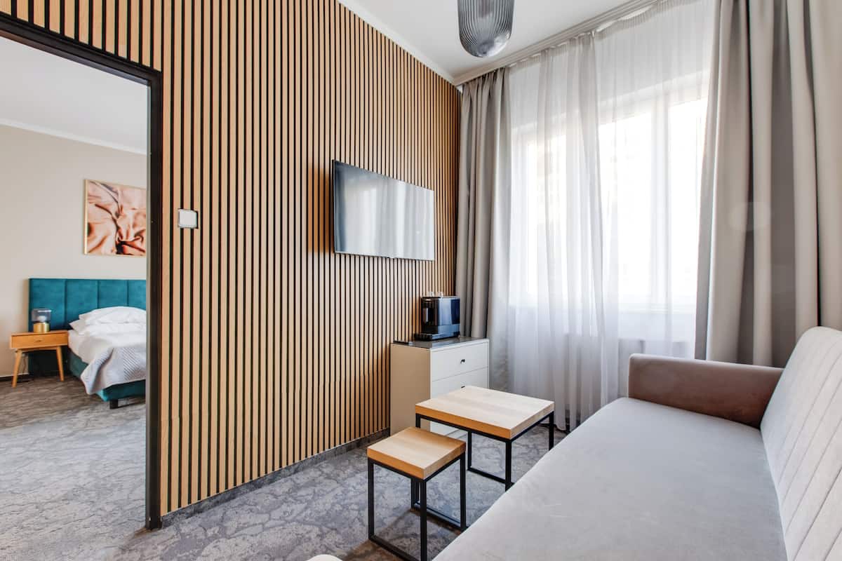 A modern hotel room with a large bed, a TV mounted on a wooden panel wall, a small table, and a view of the city through sheer curtains.