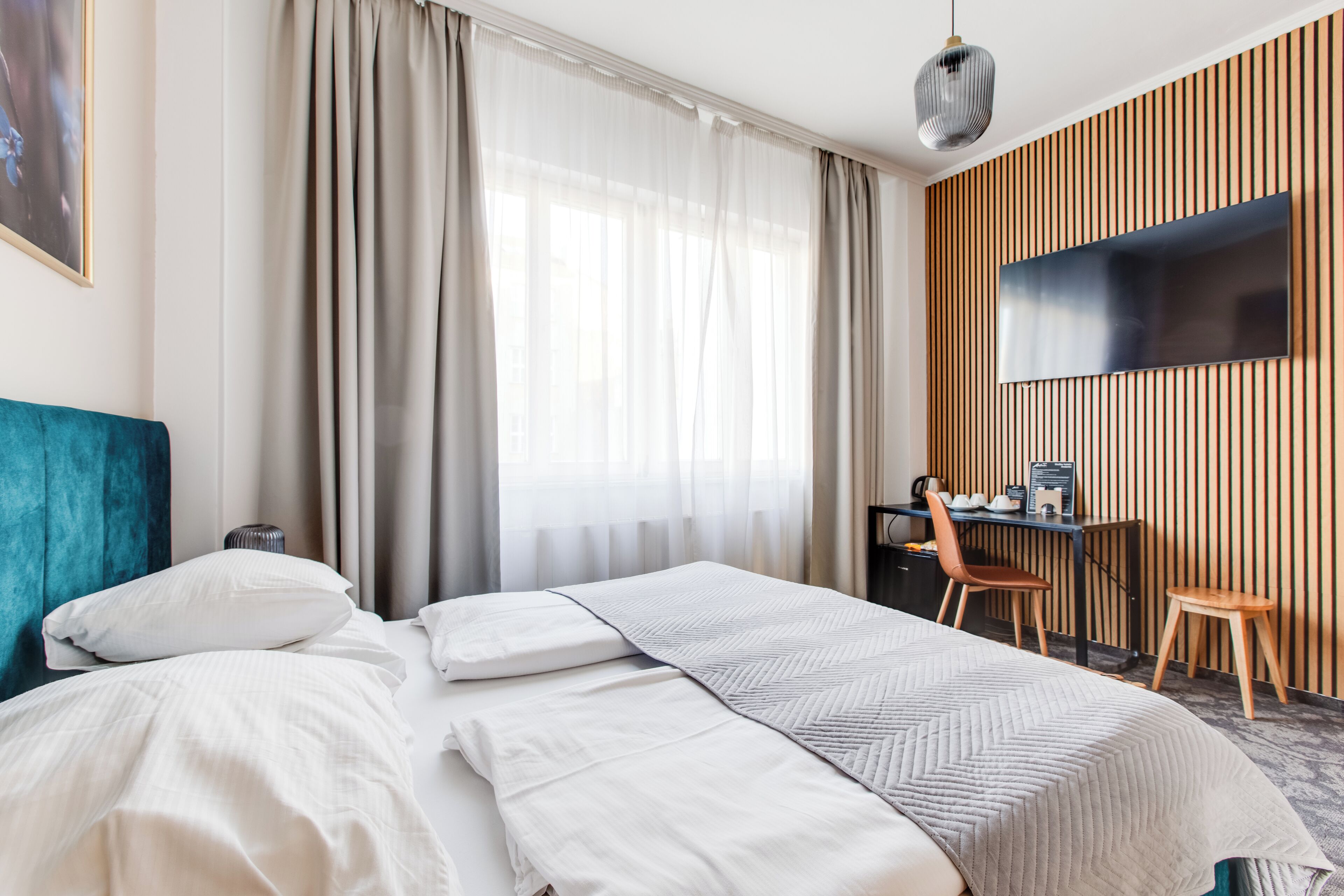 Quarto de hotel com cama, escrivaninha, cadeira, televisão e janela com cortinas.