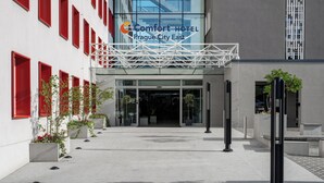 Uma entrada de hotel moderna com fachada de vidro, molduras de janela vermelhas e uma placa com o nome Comfort Hotel Prague City East.