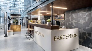 Uma área de recepção com design moderno, apresentando um balcão de atendimento identificado como "BARCEPTION", uma área de estar com cadeiras e um muro de vidro com vista para o interior.