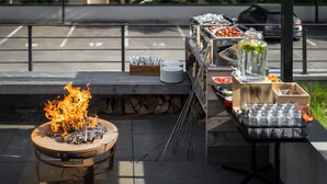 Uma área de refeições ao ar livre com lareira, mesa de buffet com diversos alimentos e uma pilha de pratos.