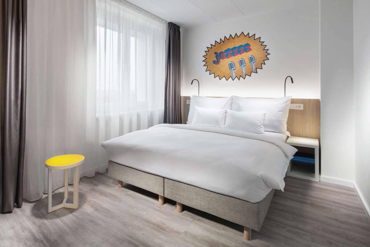 Une chambre d’hôtel moderne avec un lit, un petit tabouret jaune, une table de chevet et un élément décoratif mural.