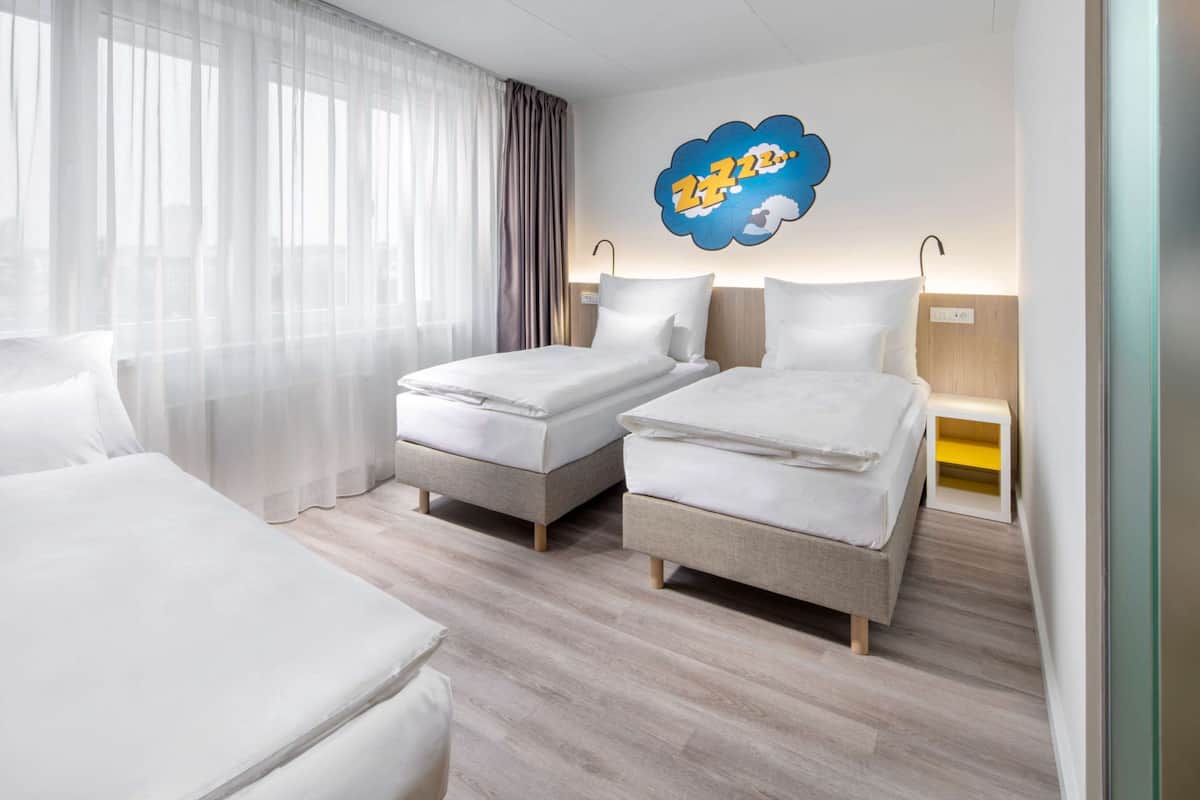 Une chambre d’hôtel avec deux lits simples, une grande fenêtre avec des rideaux légers, une œuvre d’art accrochée au mur et une table de chevet avec une lampe.