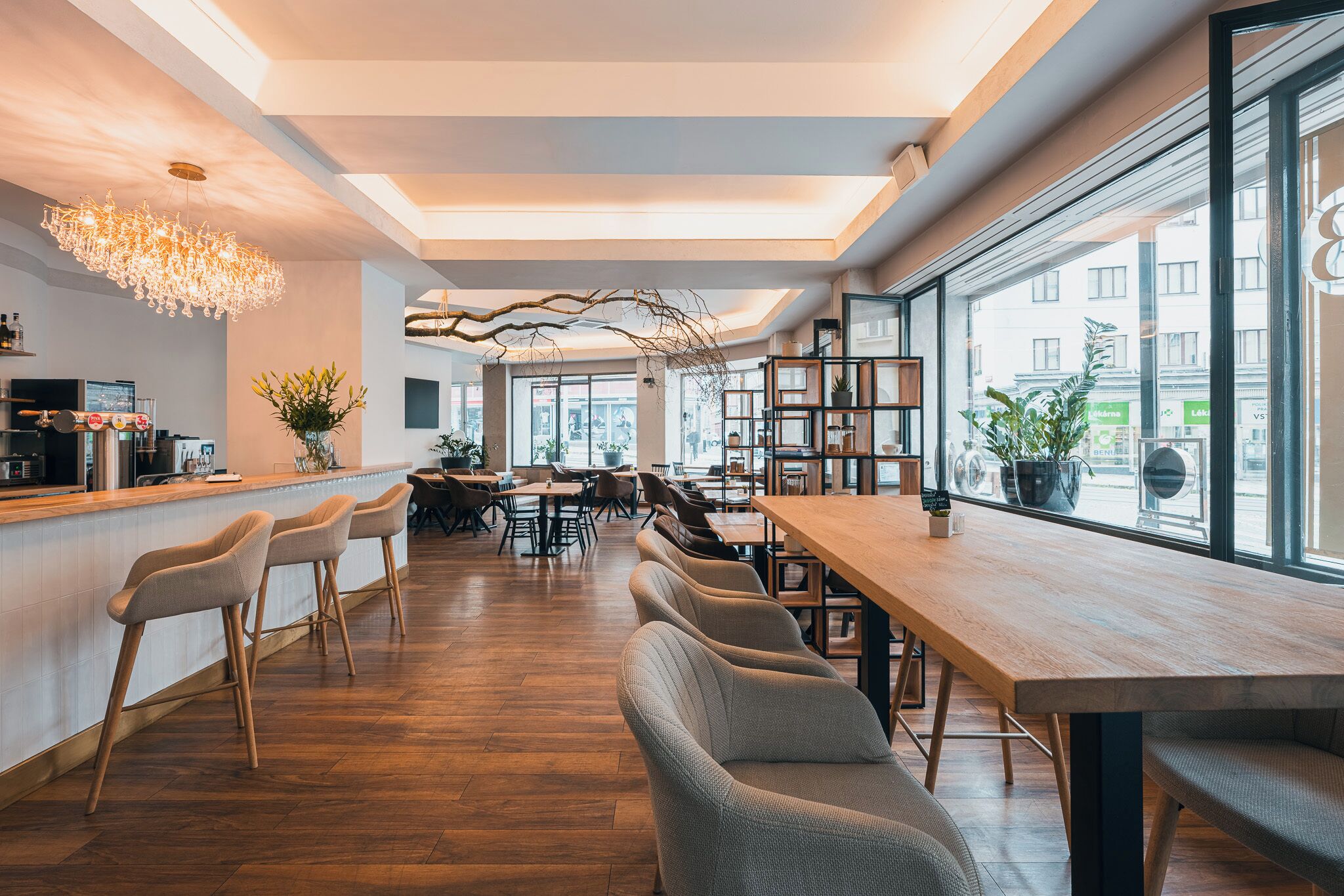 A modern cafe interior with wooden tables and chairs, a bar area, and large windows offering a view of the street.