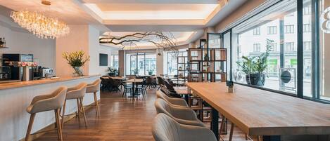 Un café moderno con mesas y sillas de madera, una barra y grandes ventanales que ofrecen vista a la calle.