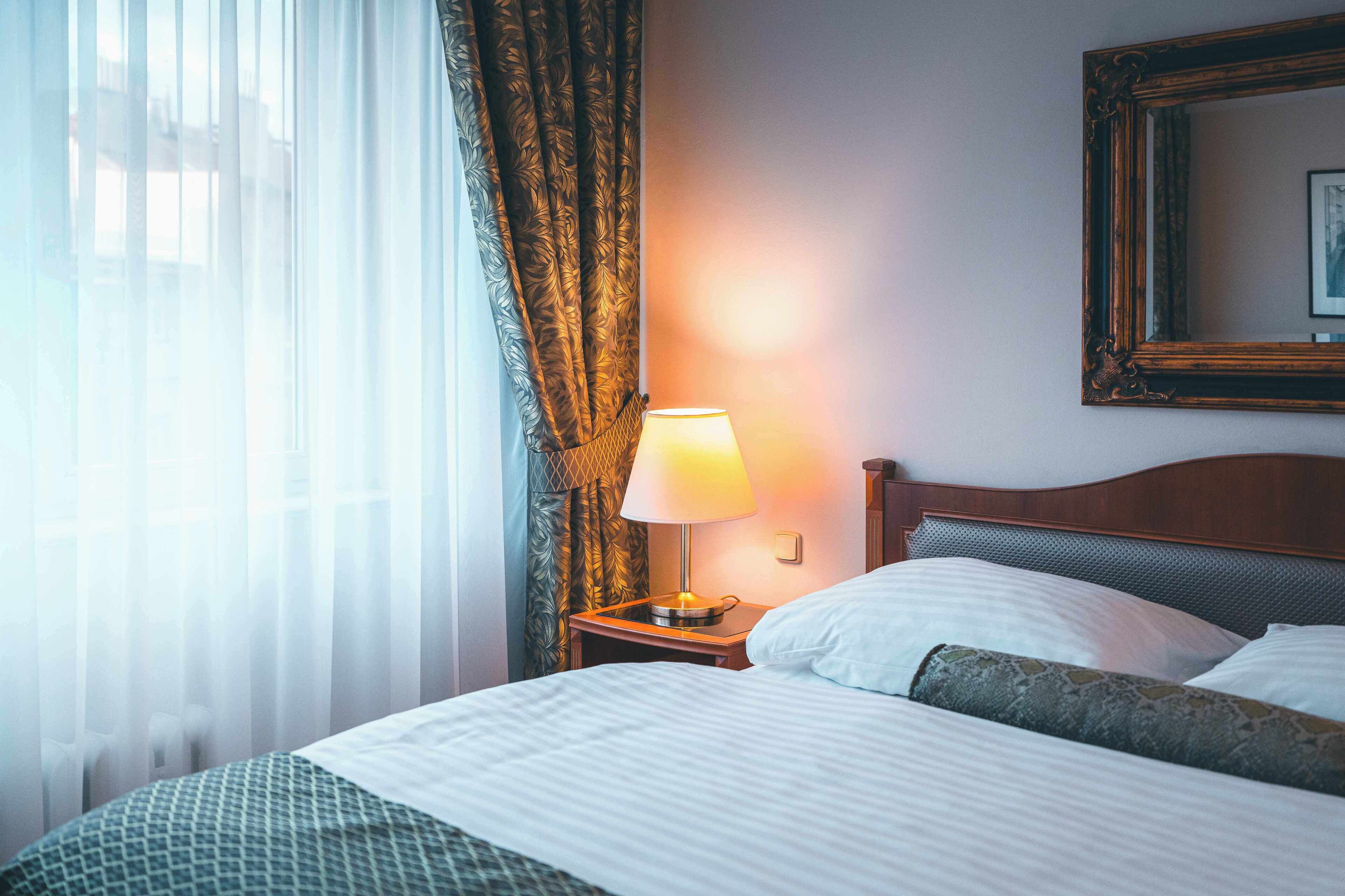 A hotel room with a bed, a nightstand with a lamp, a window with sheer curtains, and a framed picture on the wall.
