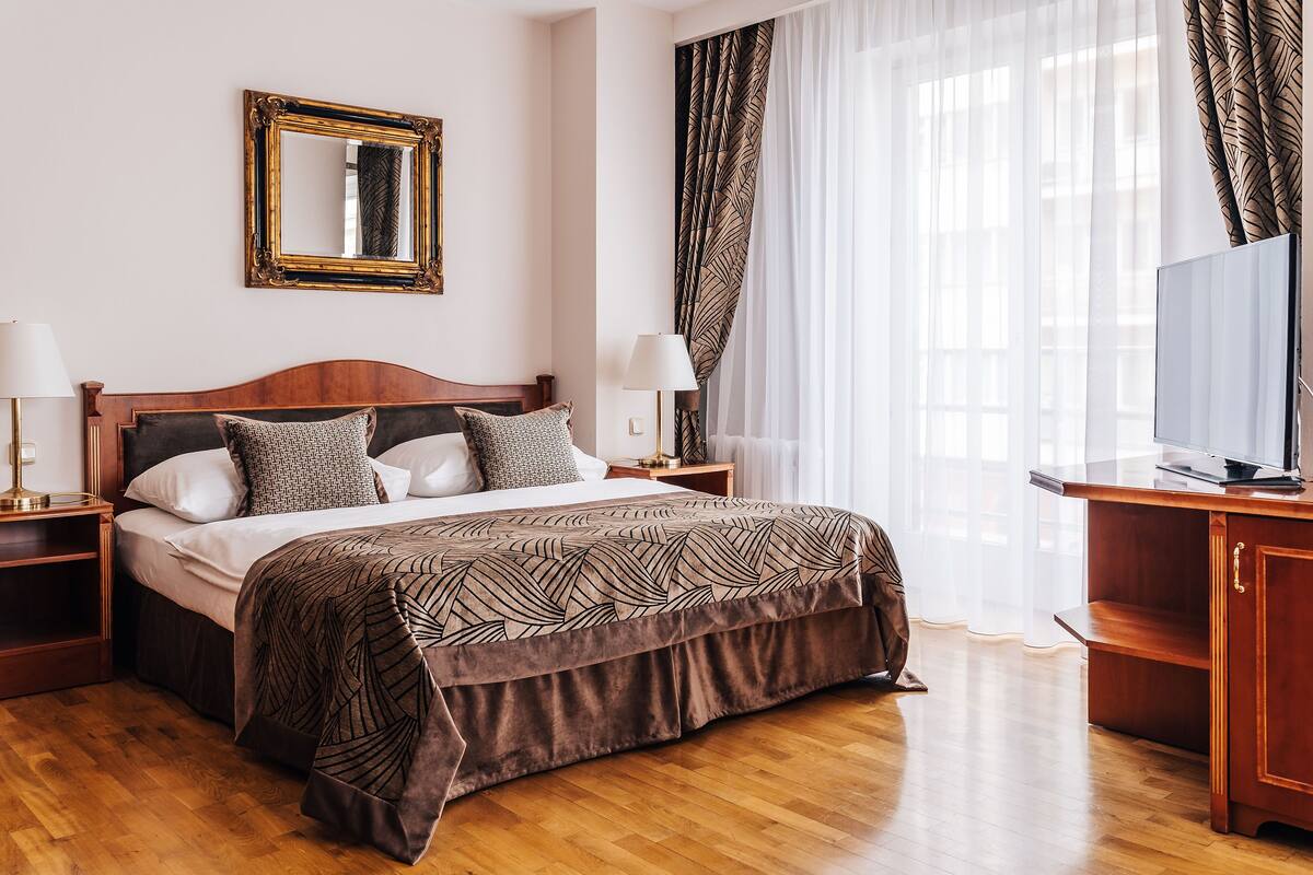 Habitación de hotel ordenada con una cama grande, dos mesitas de noche, un televisor y un amplio ventanal con cortinas translúcidas.
