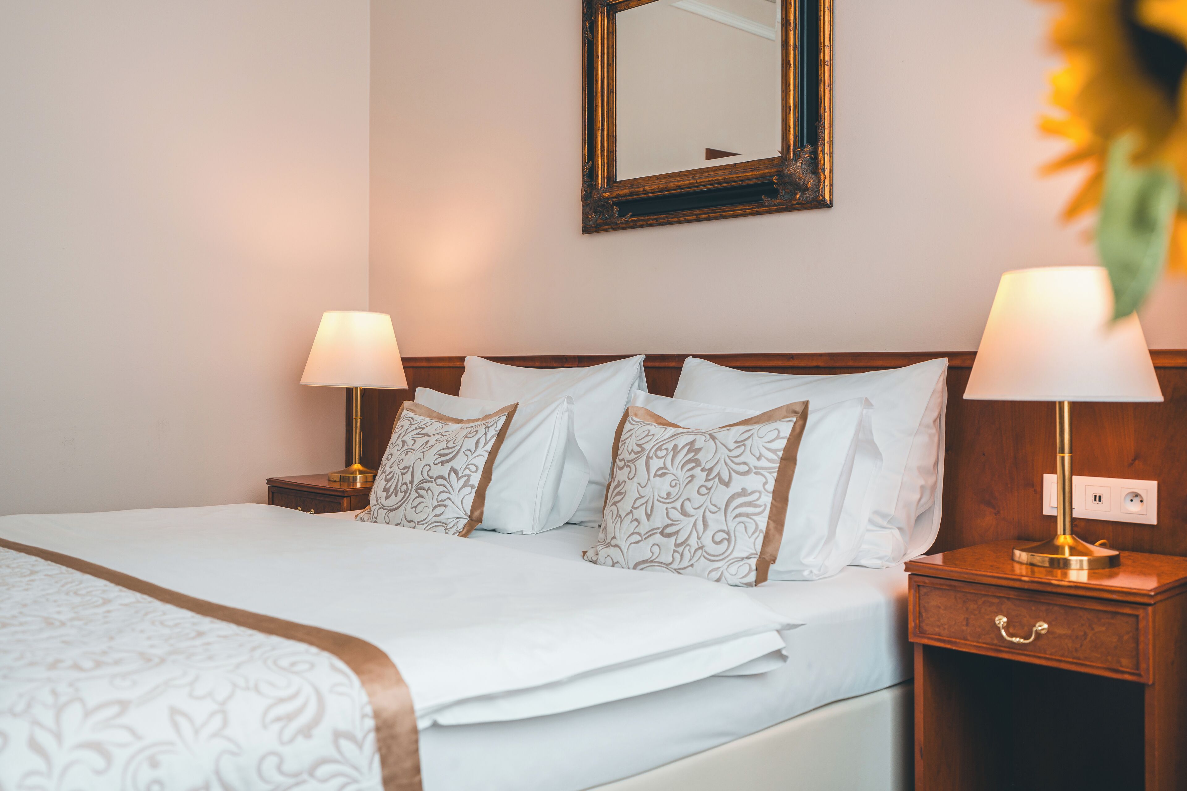 Una cama bien tendida con sábanas blancas y almohadas estampadas, dos lámparas de noche, una mesita de noche de madera y un espejo en la pared.