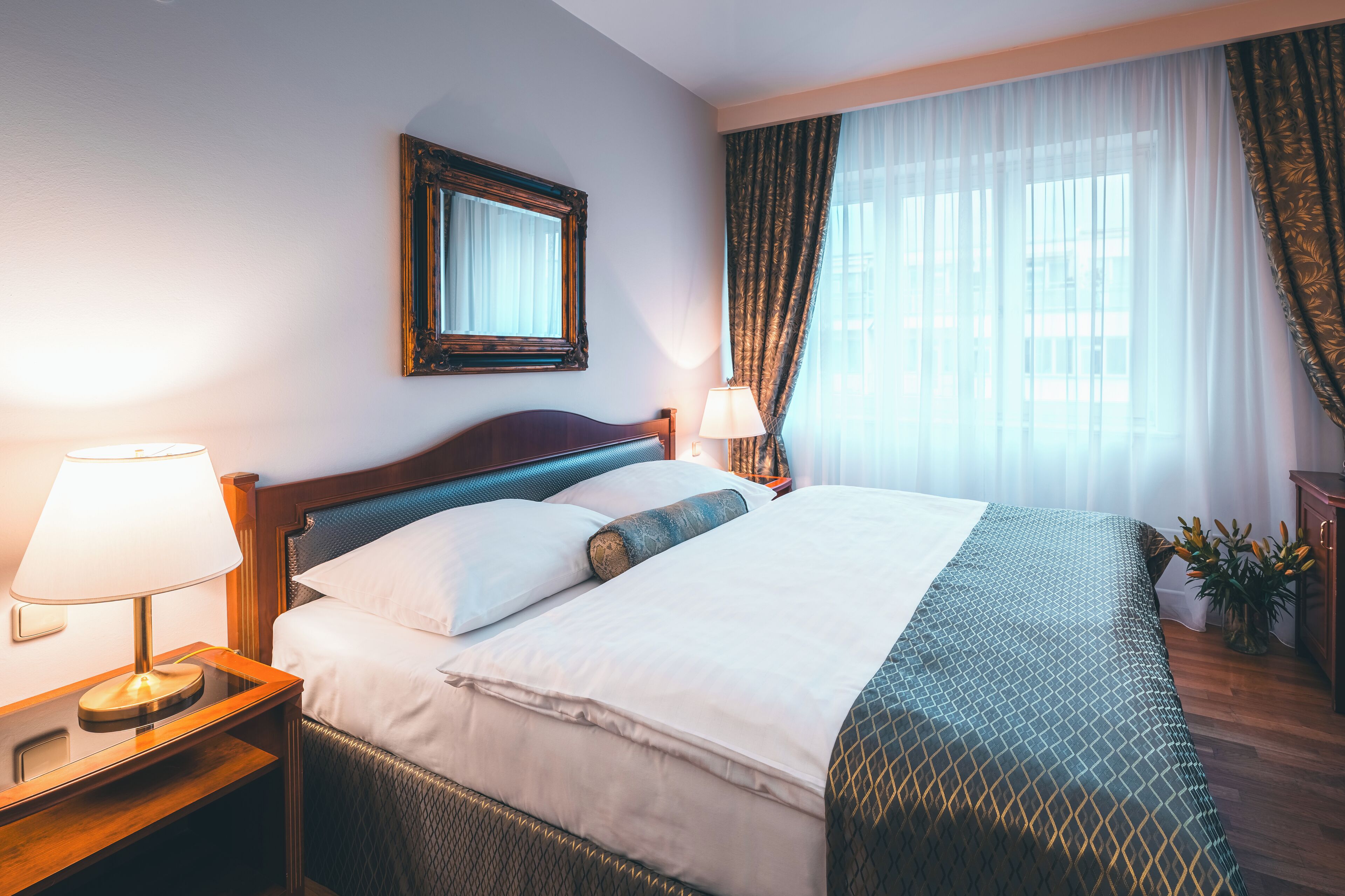 Une chambre d’hôtel avec un lit, une table de chevet avec une lampe, un miroir et une fenêtre avec des rideaux.