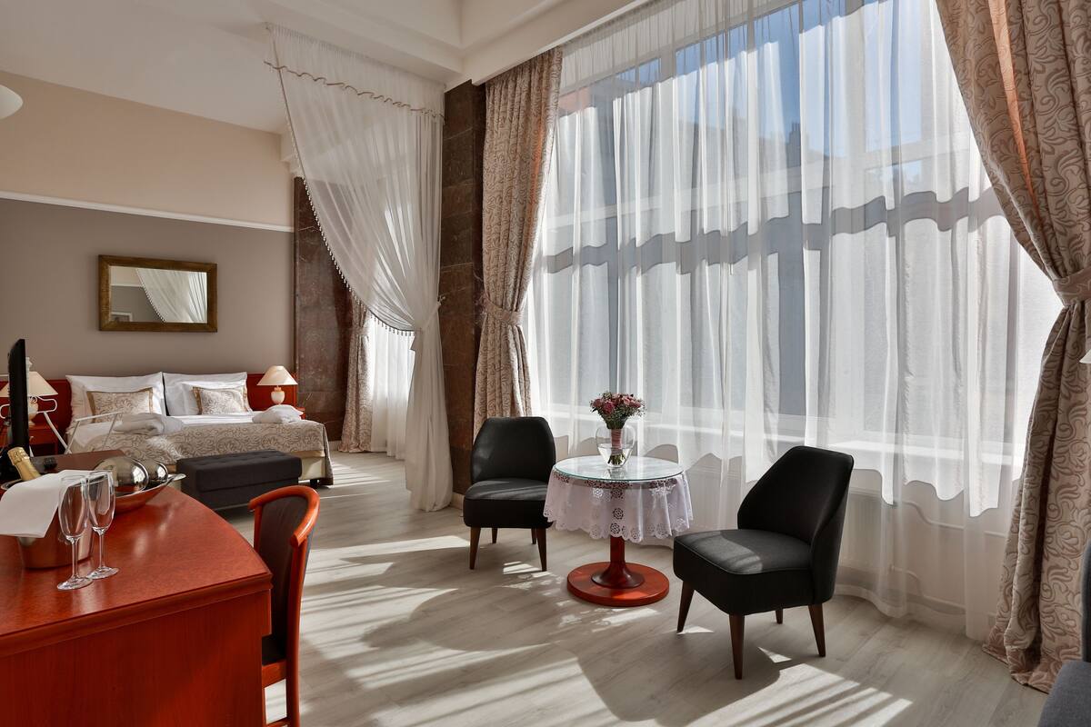 Habitación de hotel con una cama, un escritorio con dos vasos, dos sillas, una mesita con un florero y cortinas traslúcidas.
