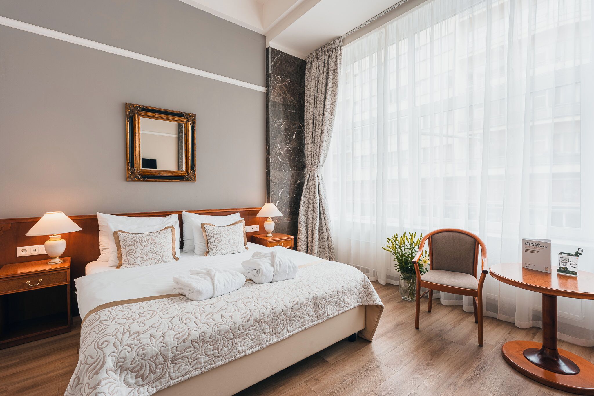 A hotel room with a large bed, two bedside tables with lamps, a chair, a small table with a sign, and a window with sheer curtains.